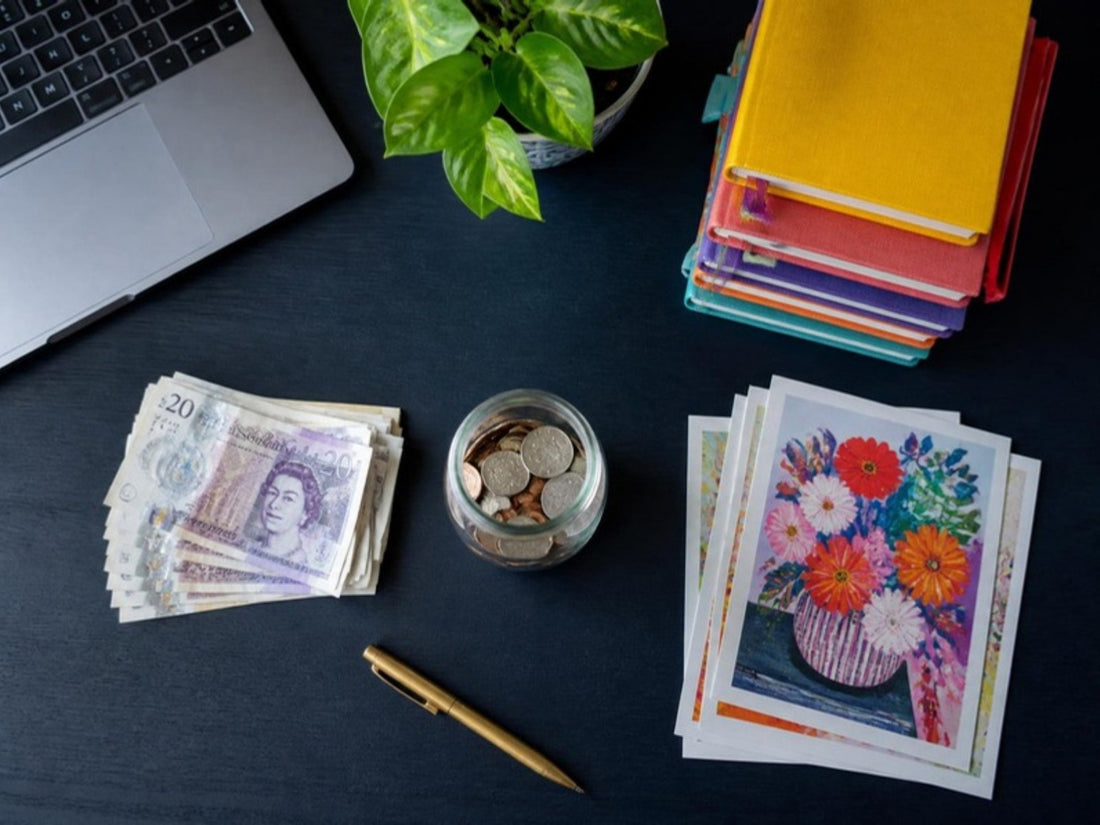 Desk with separate piles of pound notes, colourful notebooks, coins and art prints